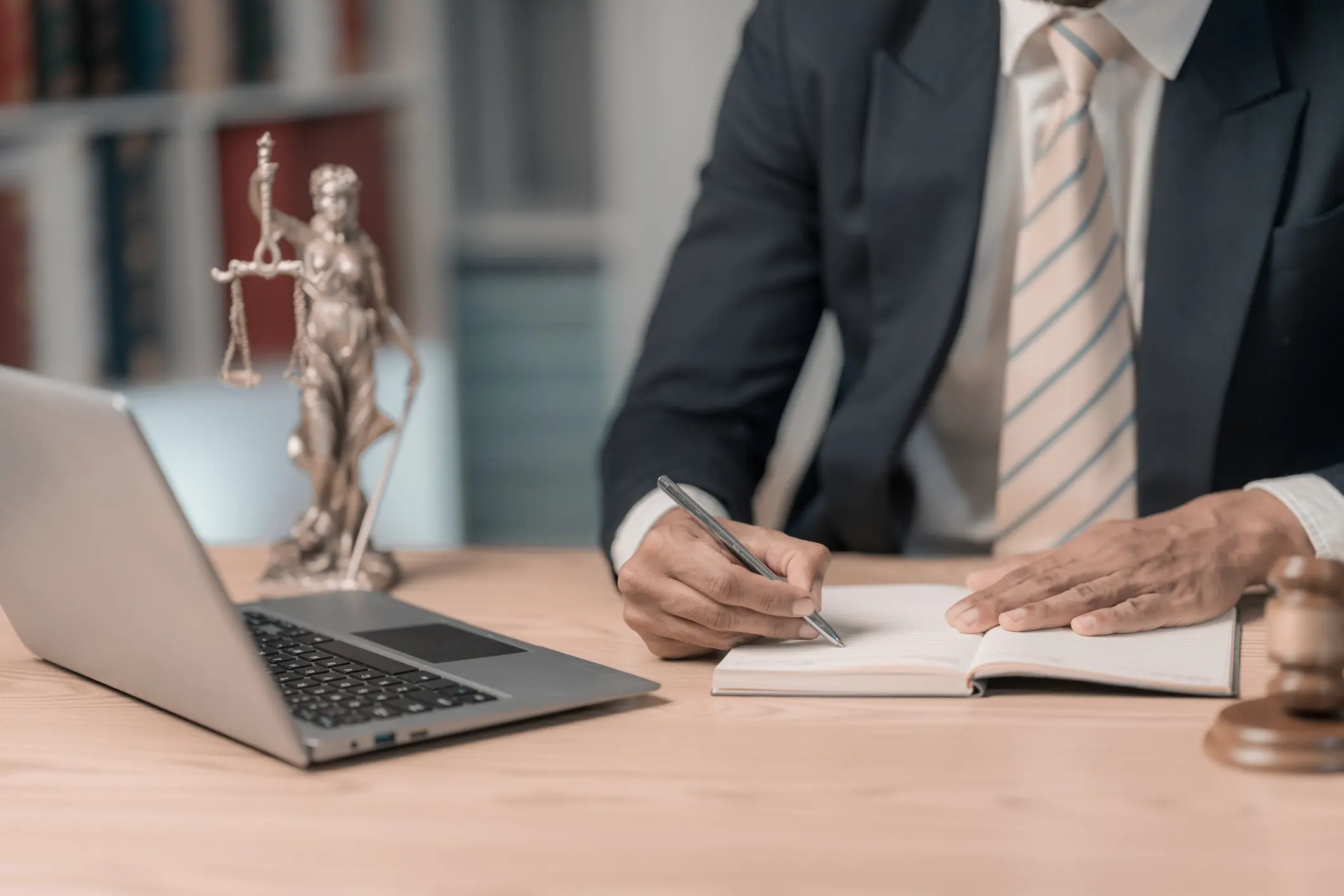 Lawyer writing notes at desk with laptop and Lady Justice statue.